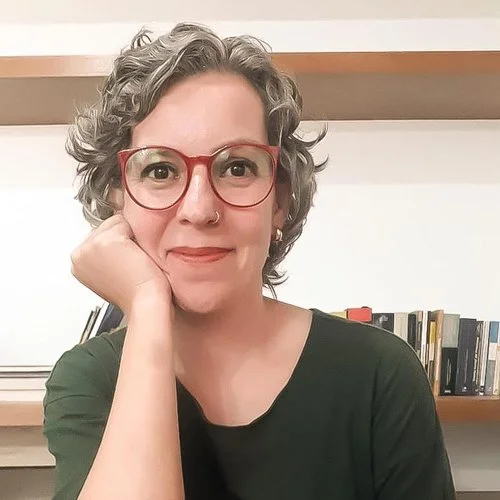 Mulher com cabelos cacheados grisalhos, óculos vermelhos, brinco de argola, sorrindo e apoiando a cabeça com a mão, em um ambiente com estantes de livros ao fundo.