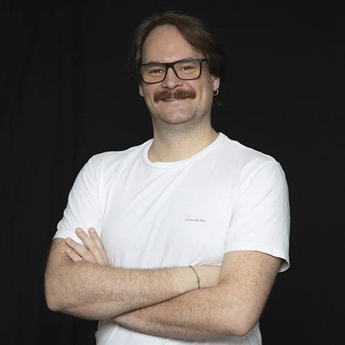 Homem com barba, óculos e cabelo castanho, sorrindo, vestindo uma camiseta branca, com os braços cruzados, contra fundo preto.