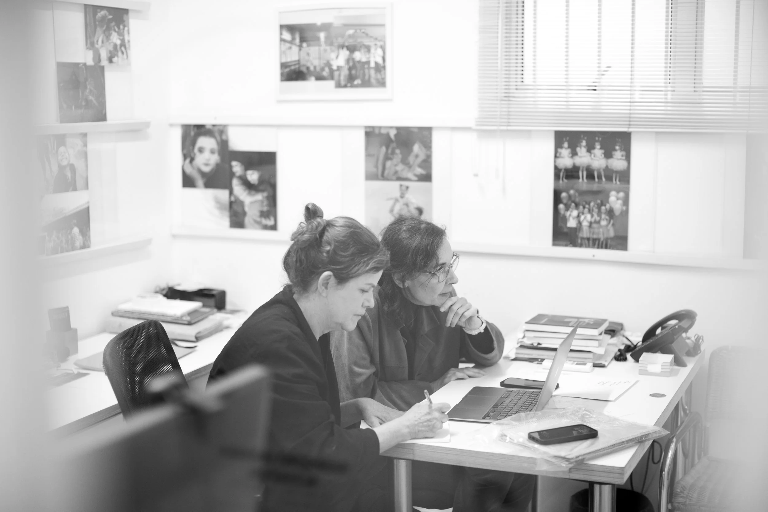 Duas mulheres trabalham juntas em uma mesa de escritório com um laptop e papéis, enquanto olham atentamente para a tela. No fundo, uma parede com fotos e uma janela com persianas.
