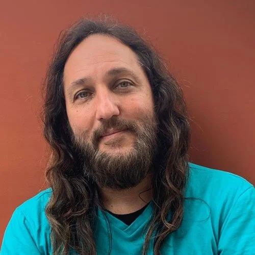 Homem de cabelo comprido, barba e bigode, usando uma camiseta azul, sorrindo levemente, com fundo laranja.