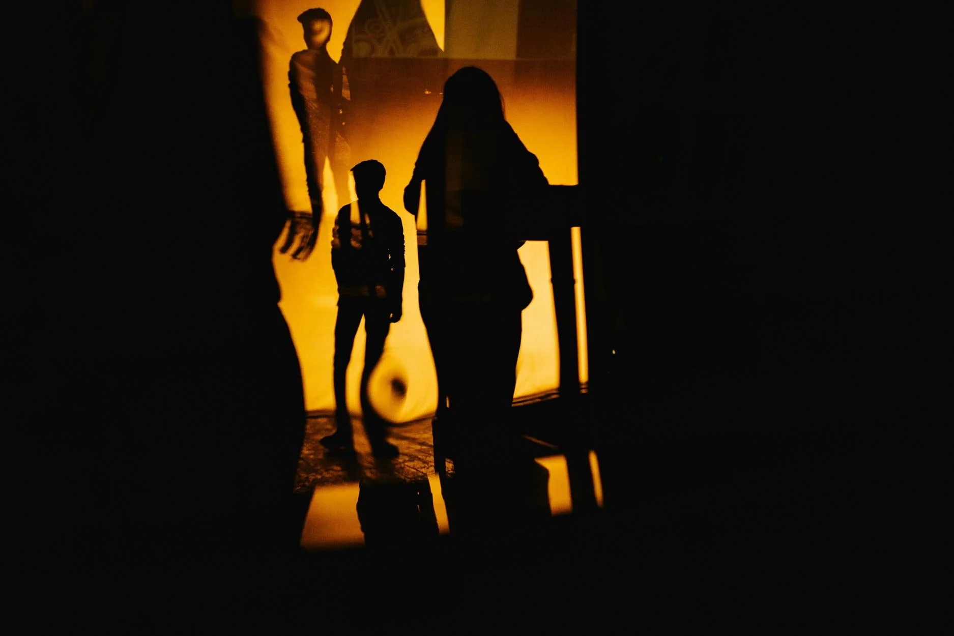 Silhueta de uma pessoa com cabelo comprido, sentada, observando uma parede de fundo iluminada por luz alaranjada, com duas outras pessoas de pé ao fundo, contraluz, em uma iluminação escura.