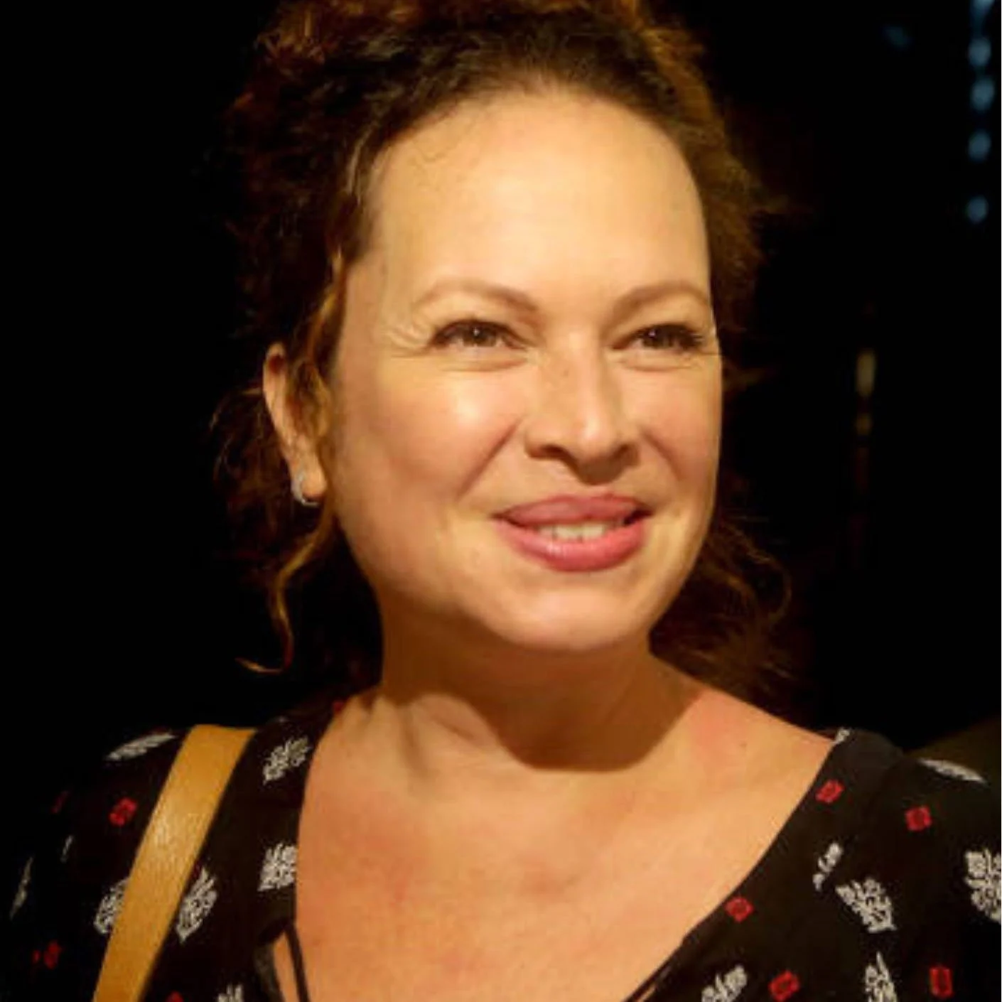 Mulher sorrindo com cabelos castanhos cacheados, vestindo blusa preta com detalhes em vermelho e branco, com fundo escuro.