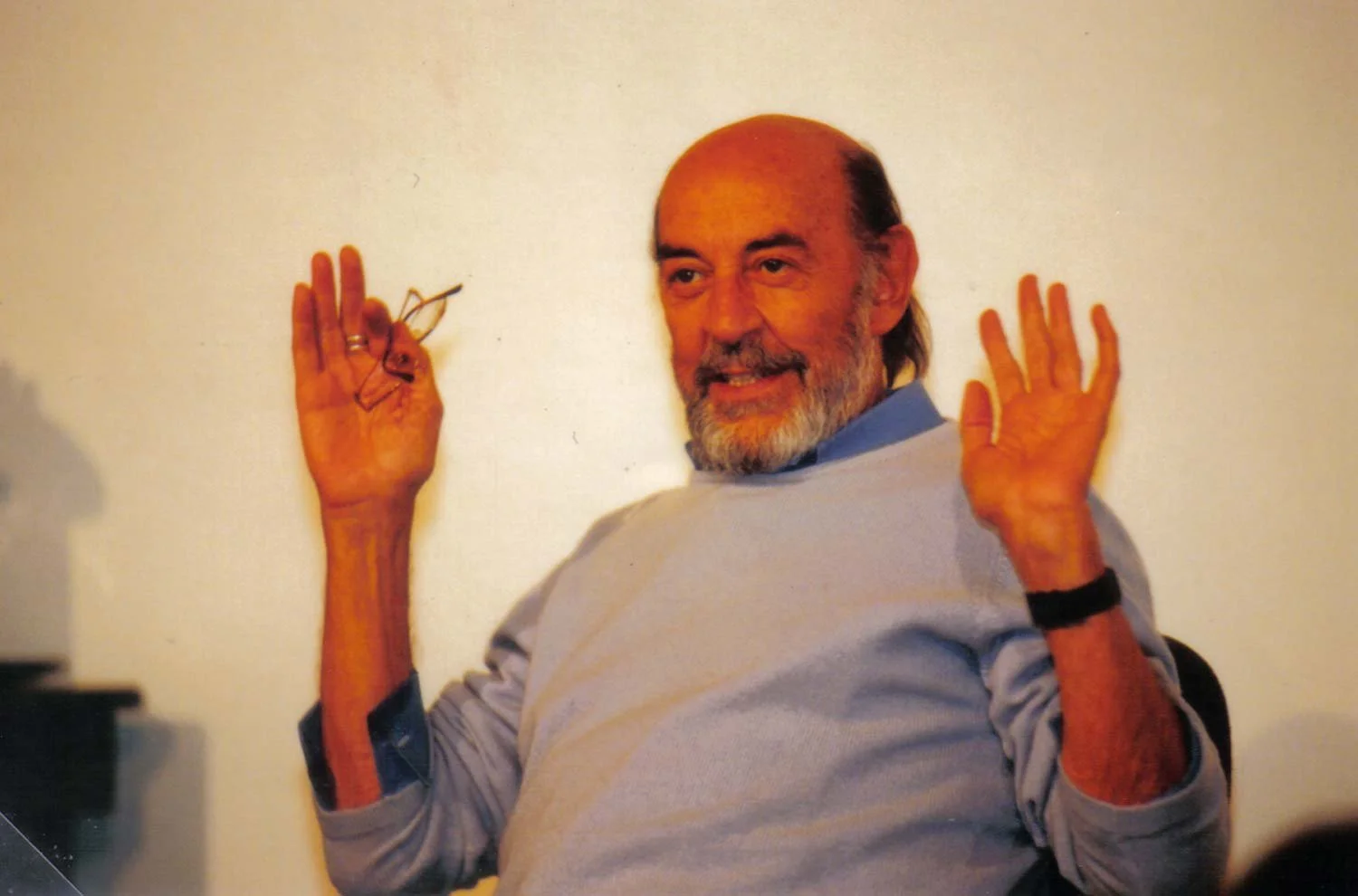 Homem sorridente com barba, vestindo suéter cinza e camisa azul, segurando um óculos na mão esquerda e fazendo um gesto de oi com a mão direita.