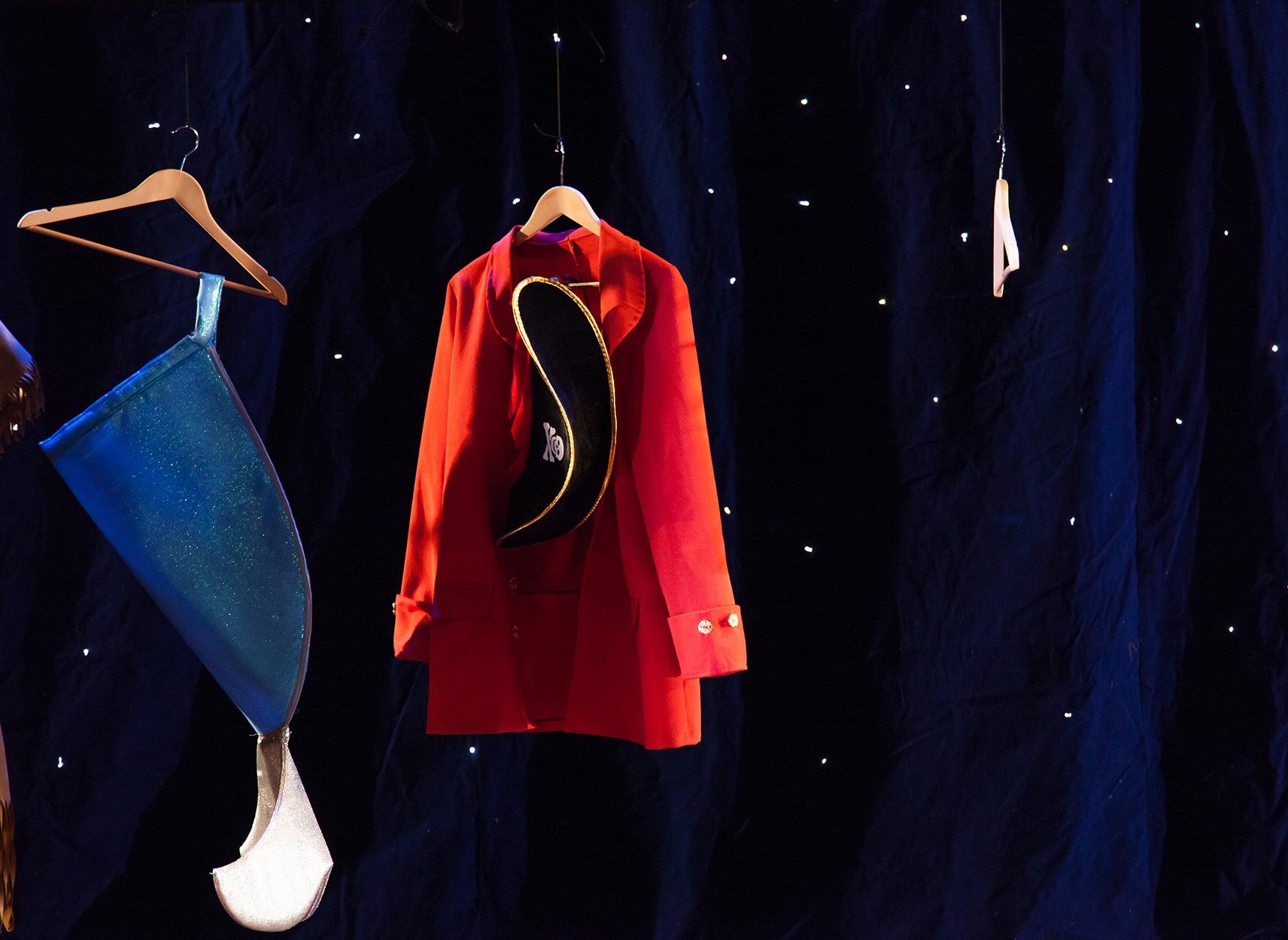 Escolha de roupas de fantasia em um fundo escuro. Há uma jaqueta vermelha, um chapéu preto com detalhes dourados e um tecido azul brilhante com glitter, além de uma meia branca de renda e uma peça de roupa branca ao longe.