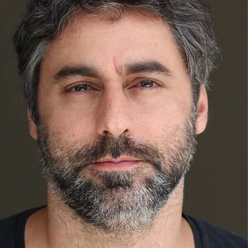 Retrato de um homem de pele clara, cabelo castanho e grisalho, barba e bigode grisalhos, olhando diretamente para a câmera, com expressão neutra, usando uma camisa preta.