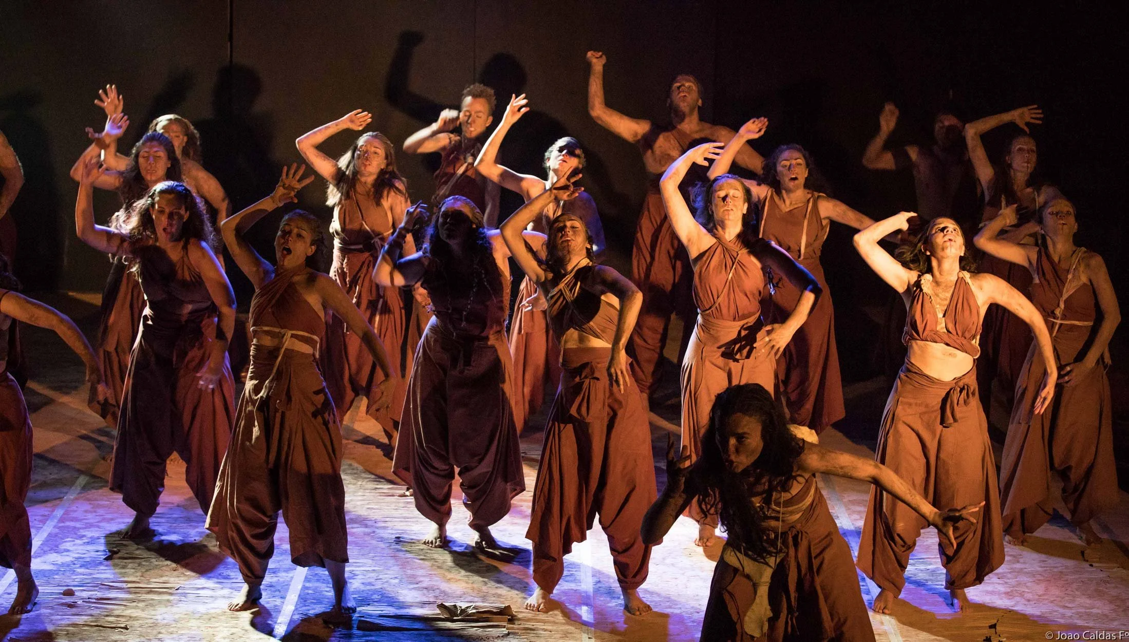 Grupo de dançarinos em palco, vestidos com roupas de cor marrom, realizando uma performance teatral em um cenário escuro com iluminação dramática.