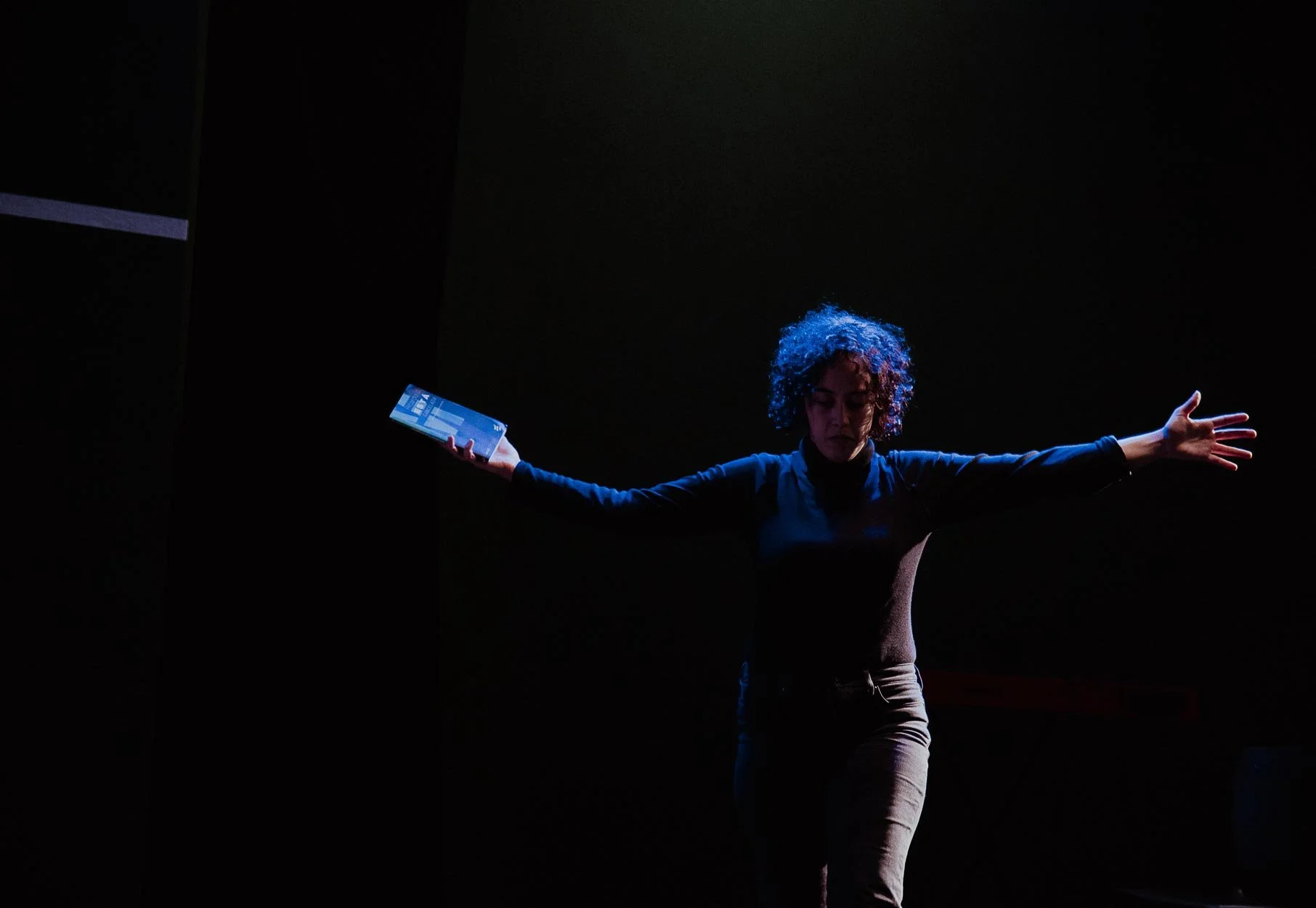 Pessoa de cabelo cacheado apoiando uma mão no ar, segurando um smartphone na outra, em fundo escuro.