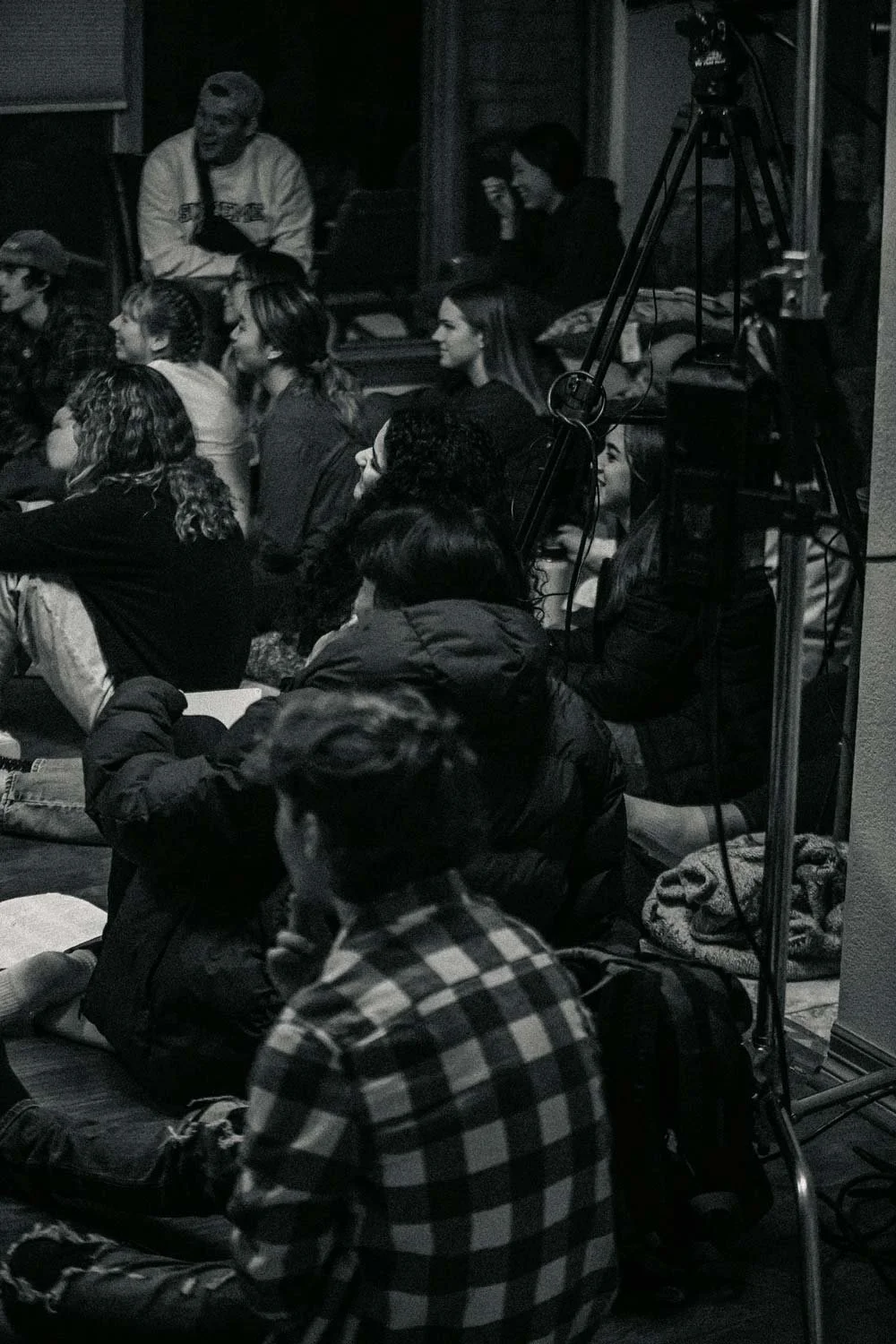 Público assistindo a uma apresentação ou palestra em um ambiente interno, com várias pessoas sentadas no chão e uma câmera de vídeo ao lado.