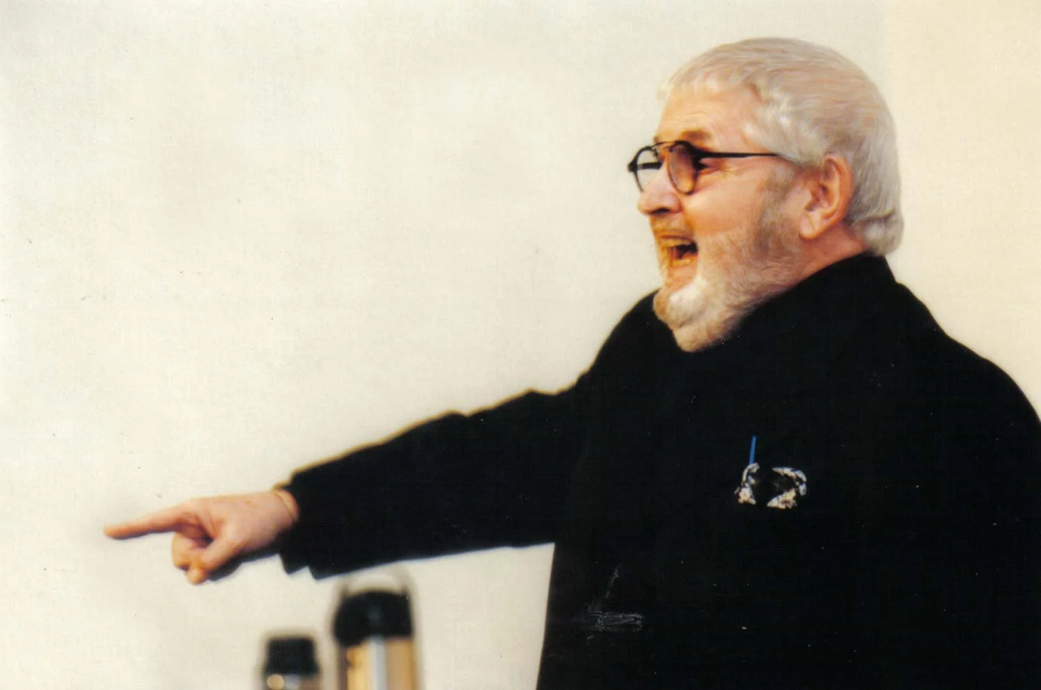 Homem idoso com cabelos grisalhos, barba e óculos, comemorando e apontando com entusiasmo, vestido de preto, ao lado de um microfone.