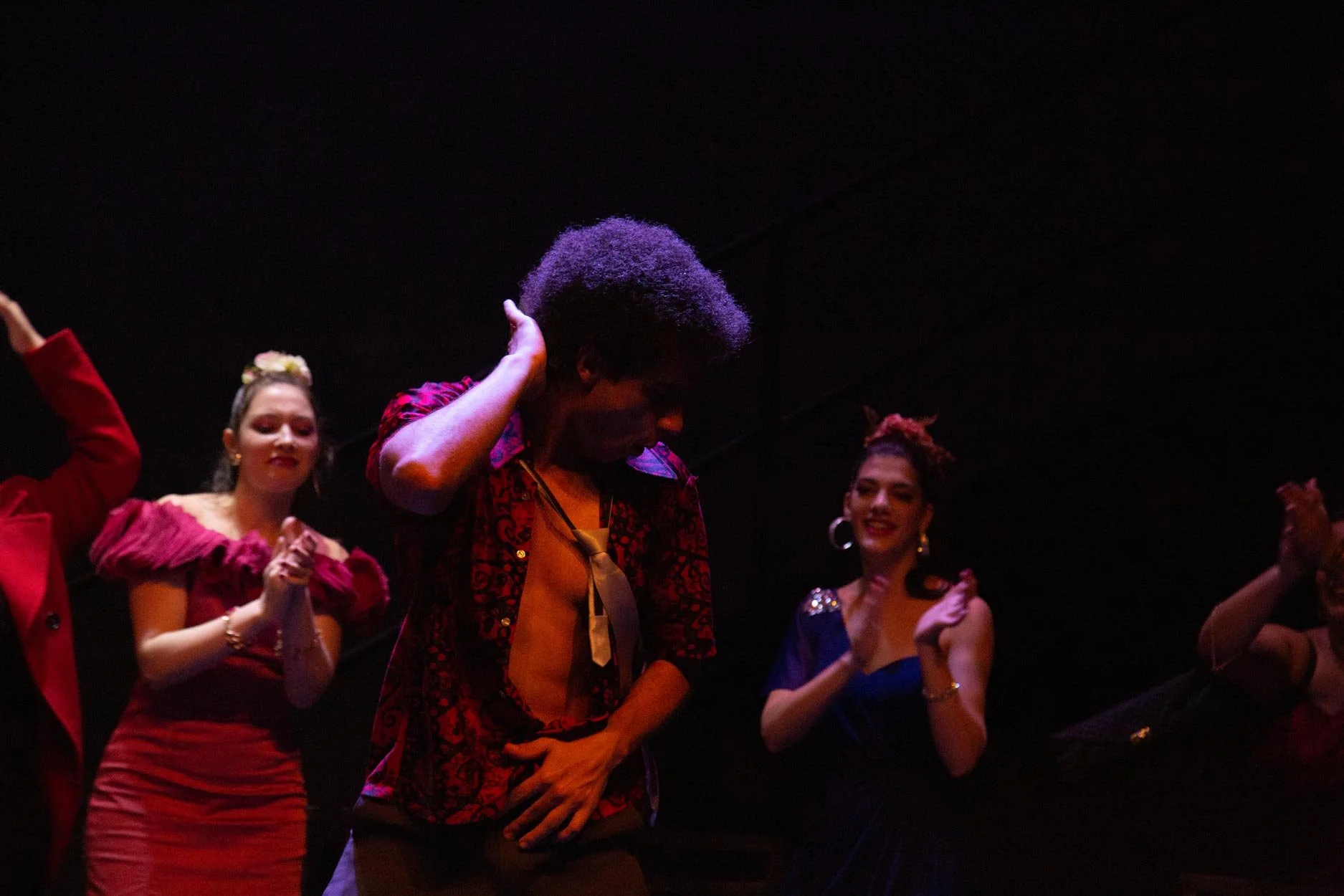 Grupo de pessoas dançando em um palco, com destaque para um homem com cabelo afro, usando uma camisa aberta e gravata, enquanto outras mulheres ao redor o aplaudem e sorriem.