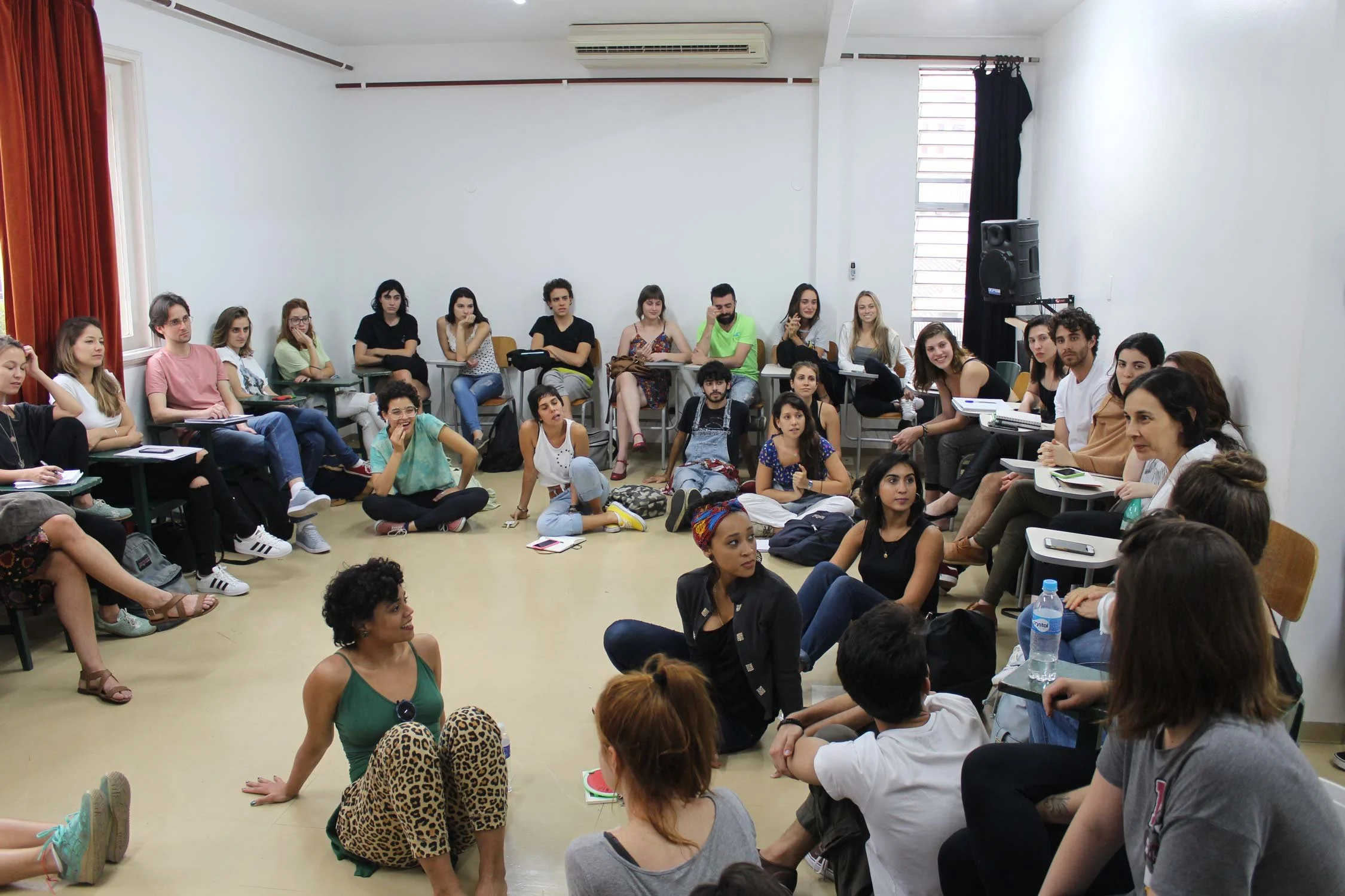 Grupo de pessoas em uma sala de aula ou reunião, algumas sentadas em cadeiras, outras no chão, participando de uma discussão ou palestra em um ambiente bem iluminado com parede branca e cortinas vermelhas e pretas.