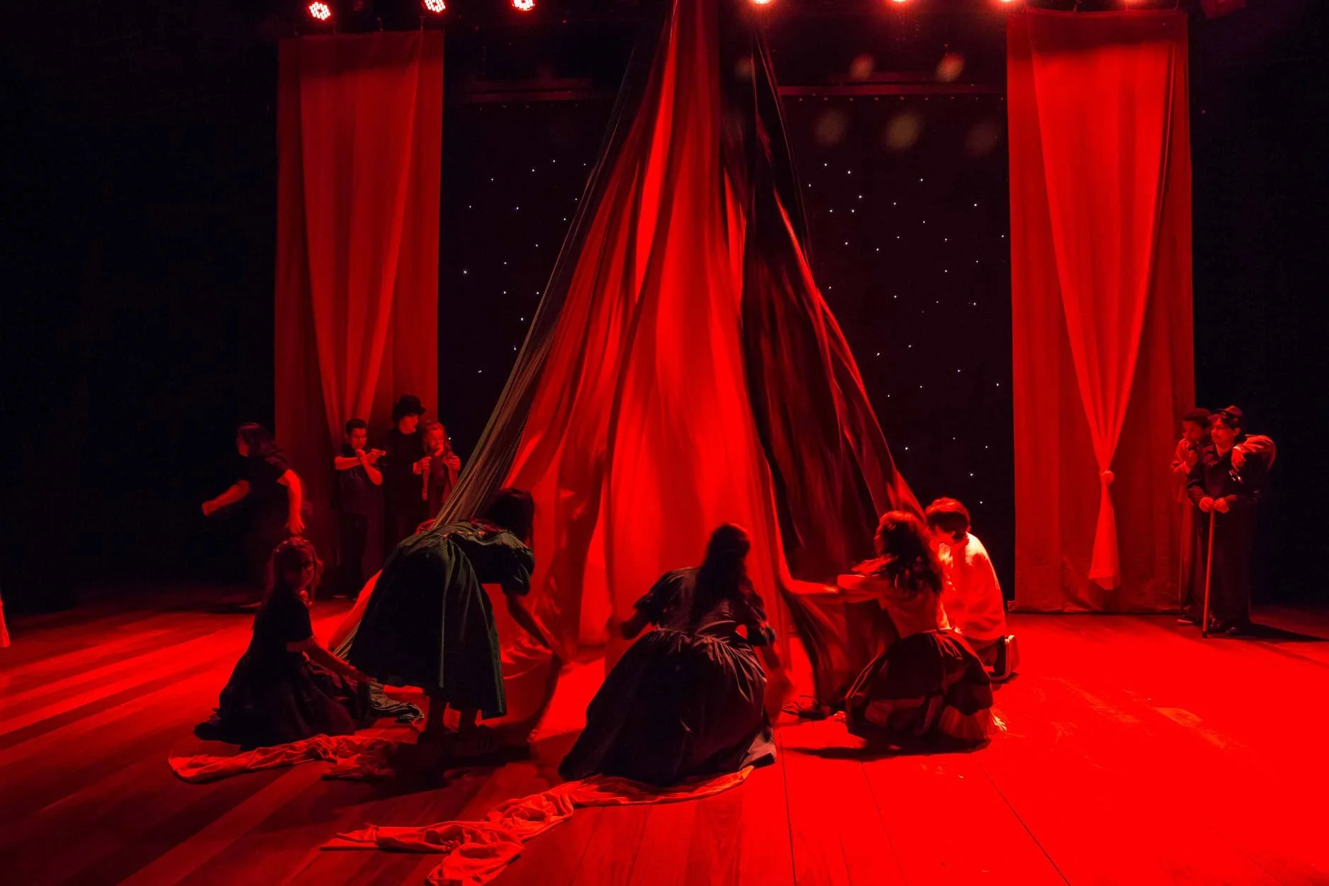 Cena de uma peça teatral com iluminação vermelha, atores com roupas antigas ao redor de um grande tecido vermelho central, com cortinas vermelhas ao fundo e estrelas no cenário.