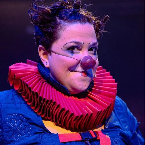 Mulher vestida de palhaça com maquiagem, cílios marcados, e rabo de cavalo, sorrindo com maquiagem de palhaço, usando figurino azul e uma gola recolorida com vermelho.
