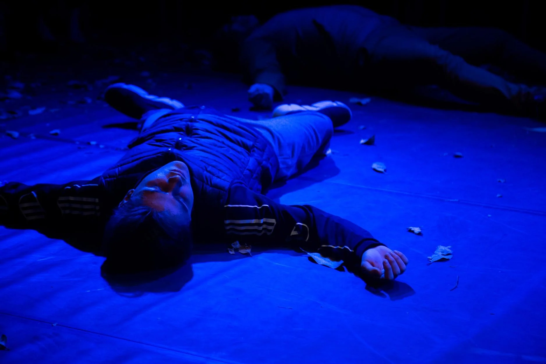 Pessoa deitada no chão sob luz azul, com folhas espalhadas ao redor, numa cena noturna ou com iluminação artificial de cores frias.