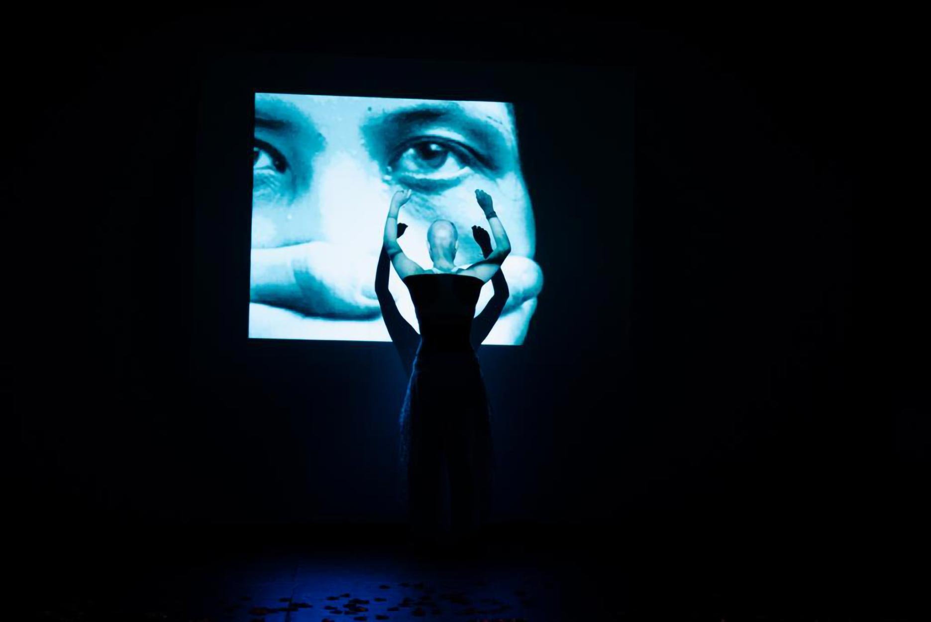 Pessoa de vestido preto de braços levantados diante de uma tela grande com imagem de rosto humano em close, numa sala escura.