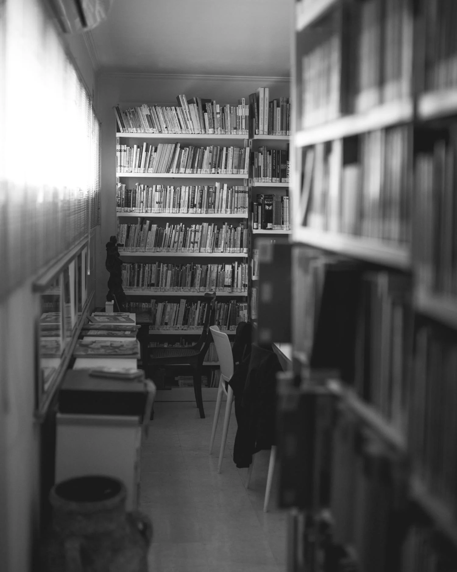 Sala de leitura com estantescheias de livros, cadeira, mesa, e iluminação natural.