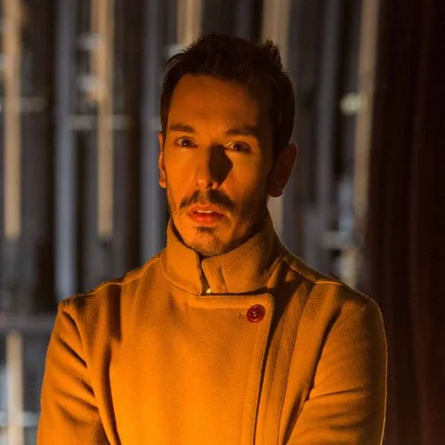 Homem com expressão séria, vestindo jaleco amarelo, em ambiente com iluminação quente e fundo parcialmente escuro.