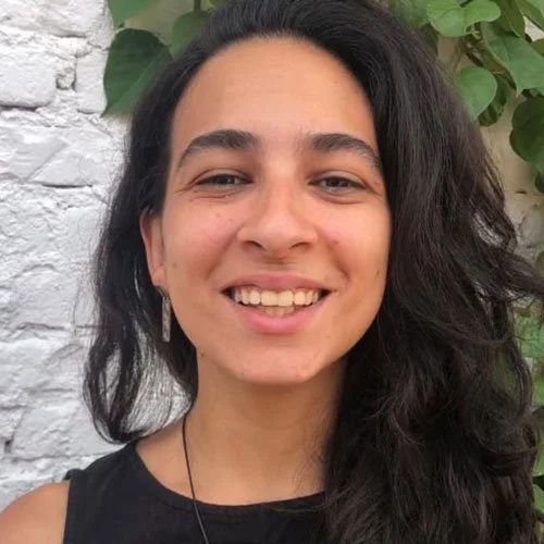 Mulher sorrindo ao lado de uma parede de tijolos brancos e plantas verdes.