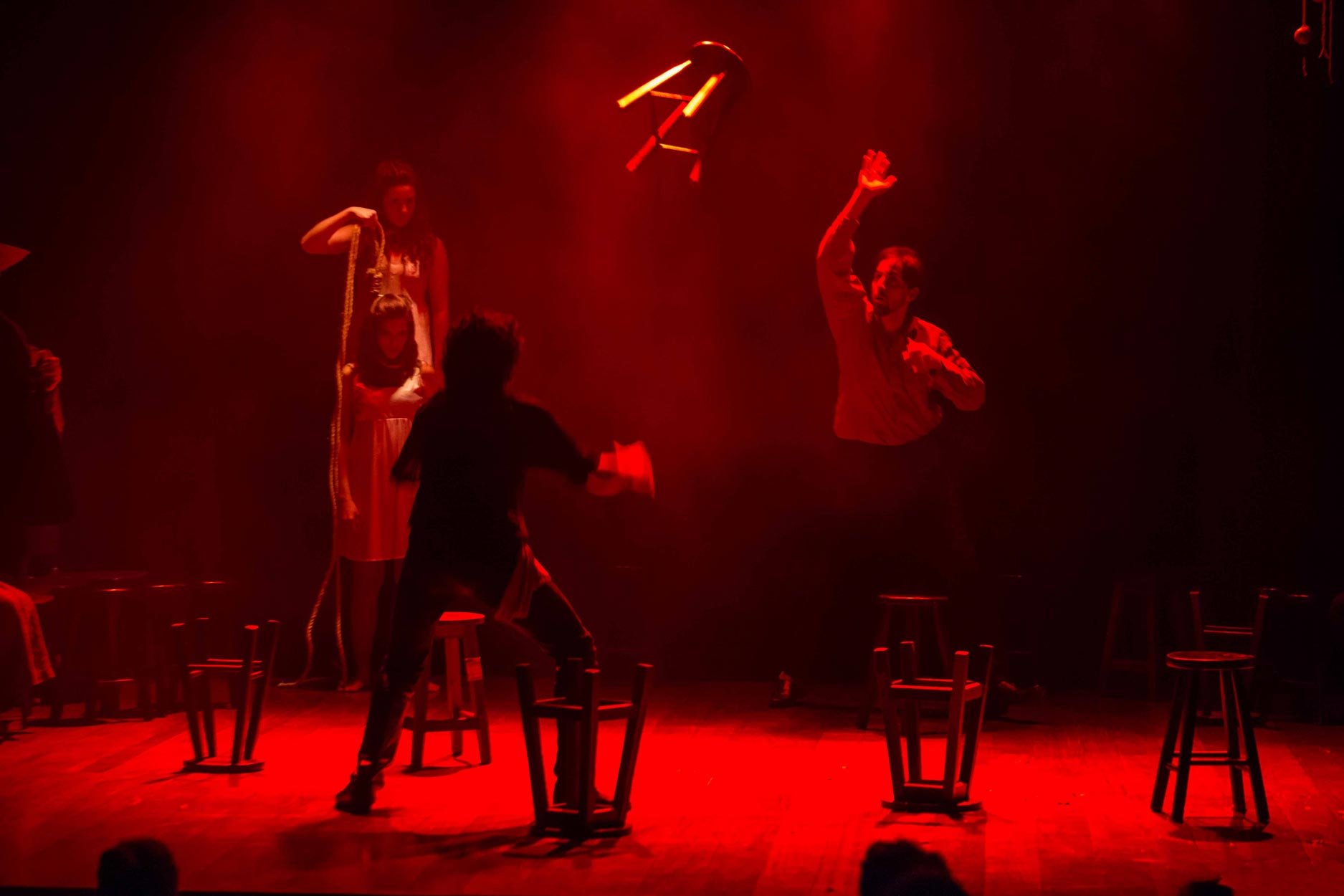 Cena de uma apresentação de teatro com iluminação avermelhada, com cinco atores no palco, alguns de pé e outros em cima de cadeiras, e objetos de teatro como cordas e utensílios suspensos no ar.