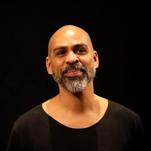 Homem com pele morena, barba grisalha, cabeça calva, usando camiseta preta, contra fundo preto.