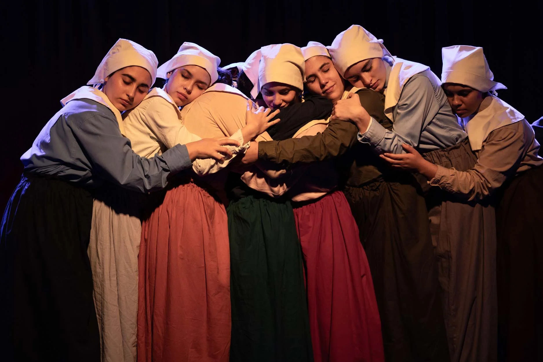 Grupo de mulheres em trajes tradicionais, com cabeças cobertas, abraçando-se em uma formação performática