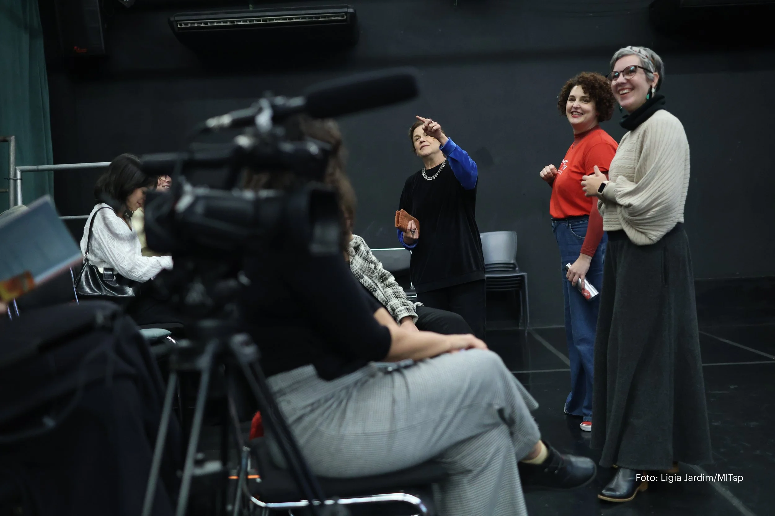 Mulheres conversando em um palco ou sala de ensaio, uma delas aponta enquanto as outras escutam, com câmera na frente registrando a cena.