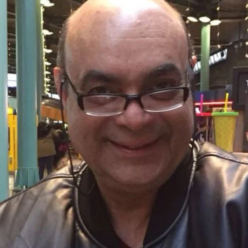 Homem sorridente usando óculos e jaqueta de couro em ambiente de transporte público ou estação de trem, com iluminação artificial e algumas pessoas ao fundo.