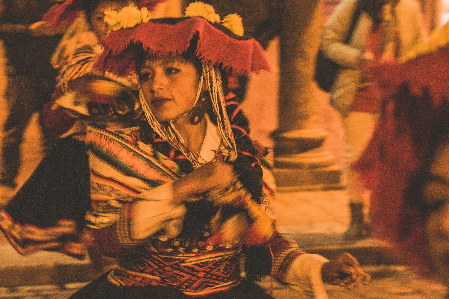 Mulher indígena em trajes tradicionais dançando, usando um chapéu decorado com penas e colares, em ambiente interno