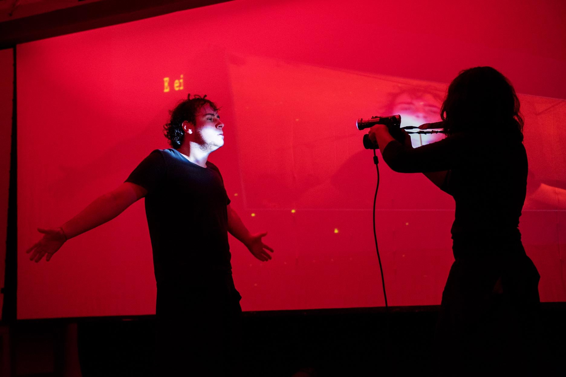 Pessoa com cabelo cacheado, de camiseta preta, posando com braços abertos diante de uma tela vermelha, enquanto outra pessoa com cabelo longo e escuro aponta uma arma de brinquedo na direção dela, em um ambiente com iluminação baixa.