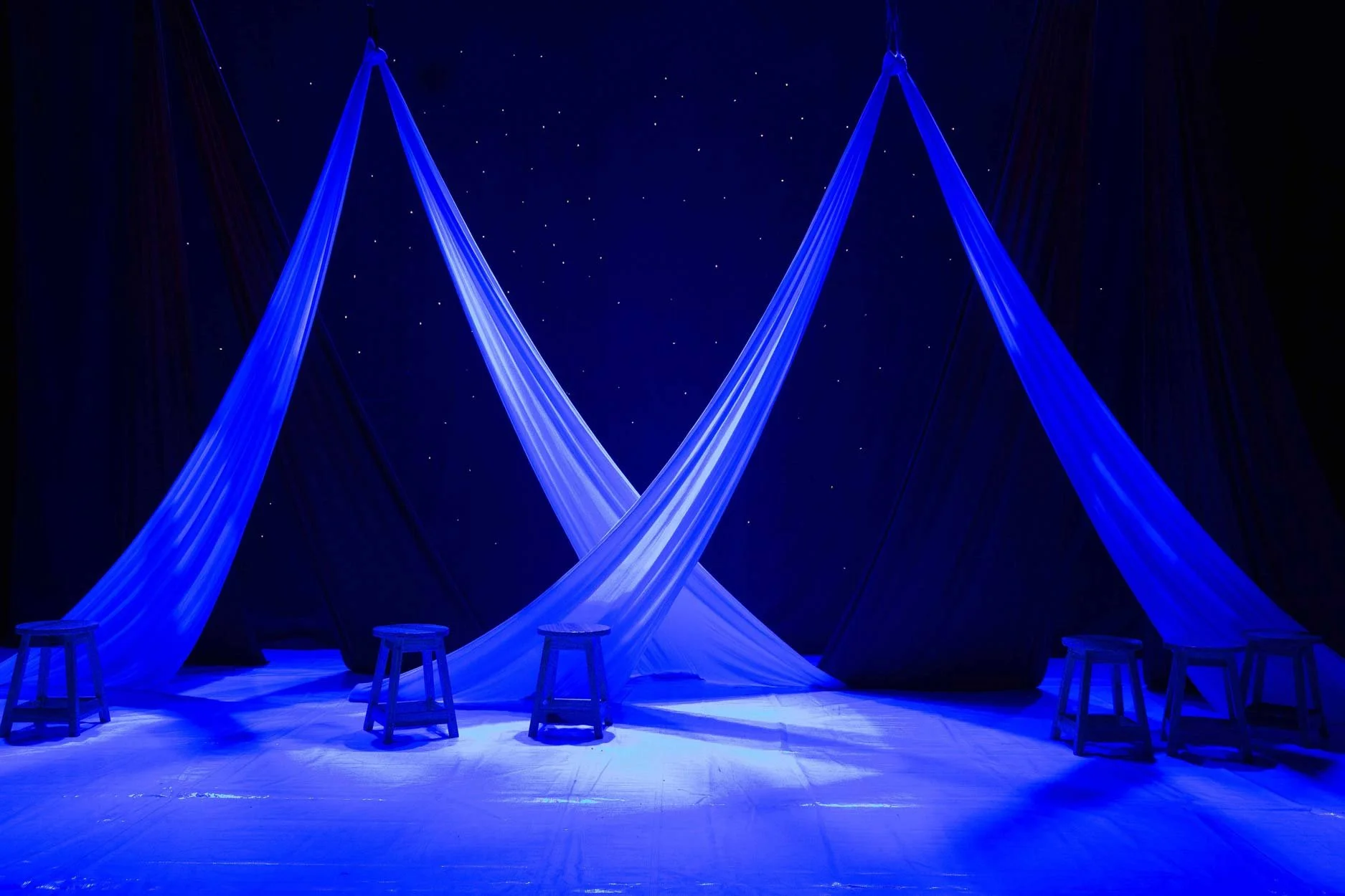 Cortina azul e preta com cadeiras de madeira e um espaço escuro no fundo com estrelas.