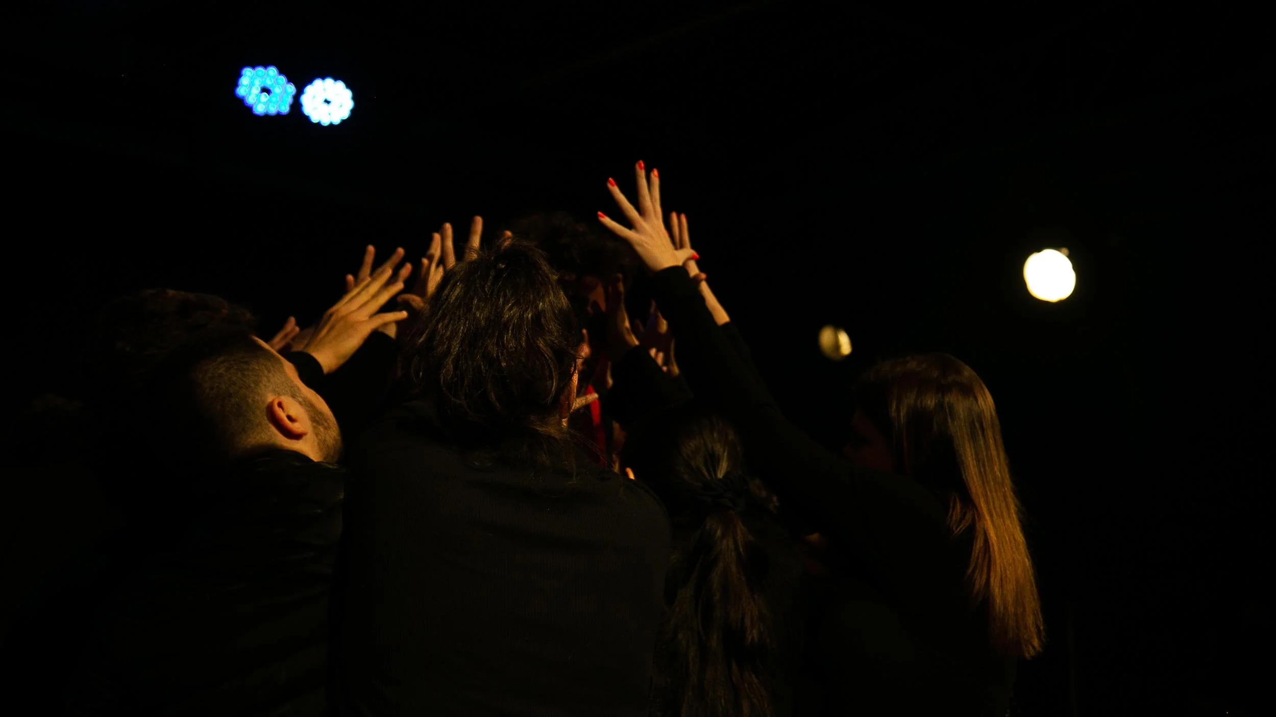 Grupo de pessoas com as mãos levantadas durante uma apresentação ou evento em ambiente escuro com iluminação baixa.