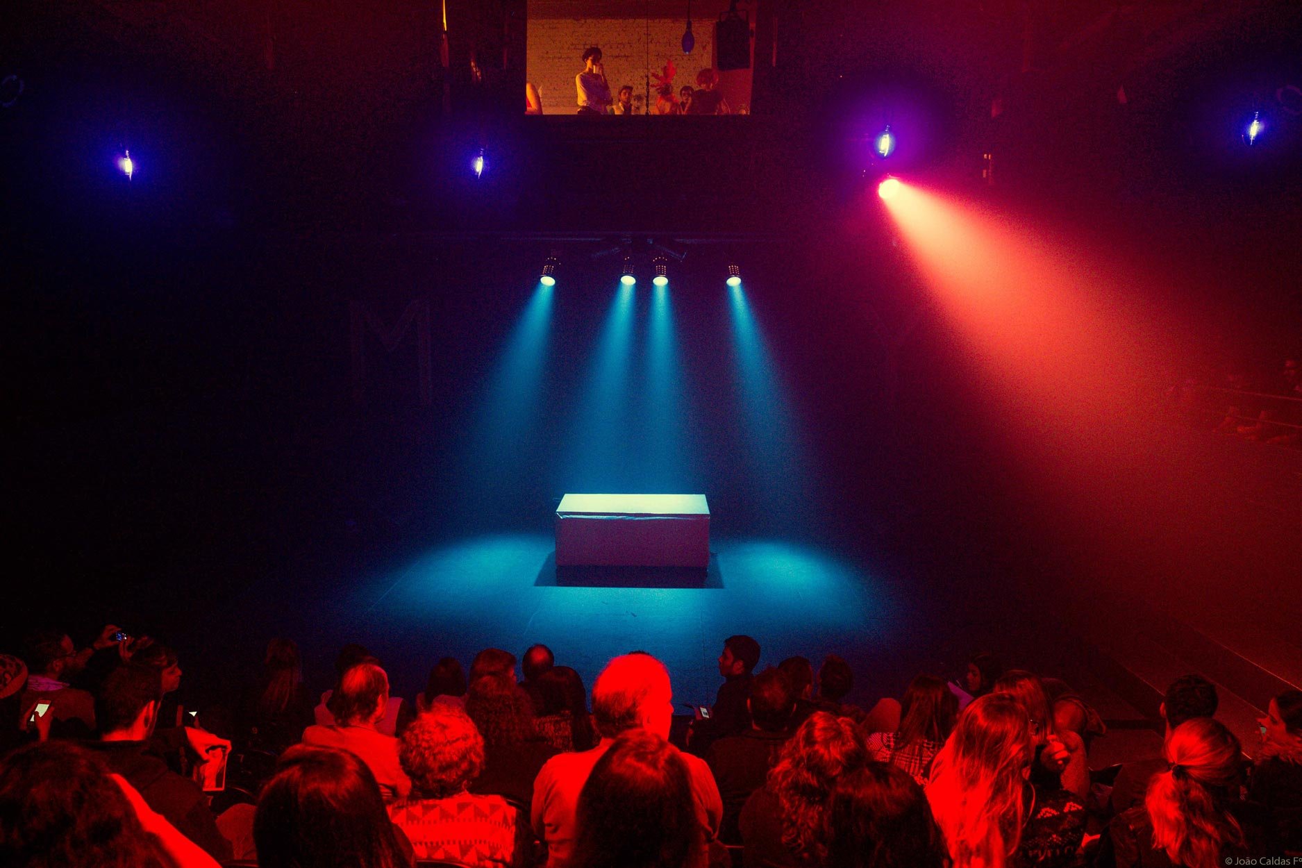 Cena de um palco de teatro ou cinema com iluminação azul e vermelha, uma mesa no centro e espectadores assistindo ao espetáculo.
