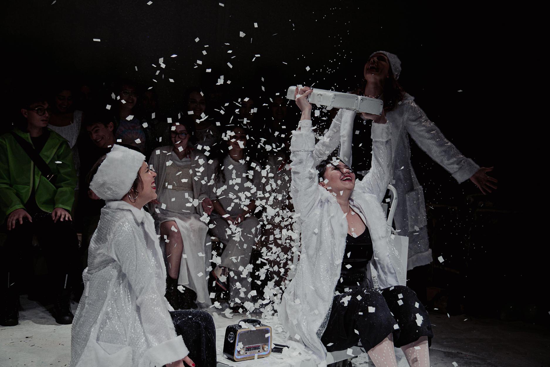 Mulheres comemorando uma conquista, uma delas com um presente na cabeça, enquanto outras assistem e sorriem ao redor, com confetes caindo e um cenário escuro.