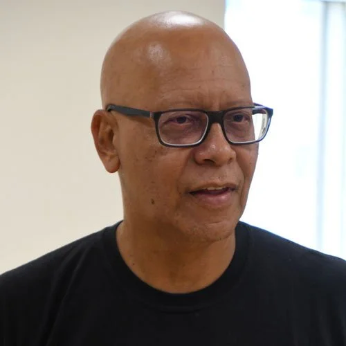 Homem com pele clara, cabelo pelado, usando óculos e camiseta preta, em ambiente interno