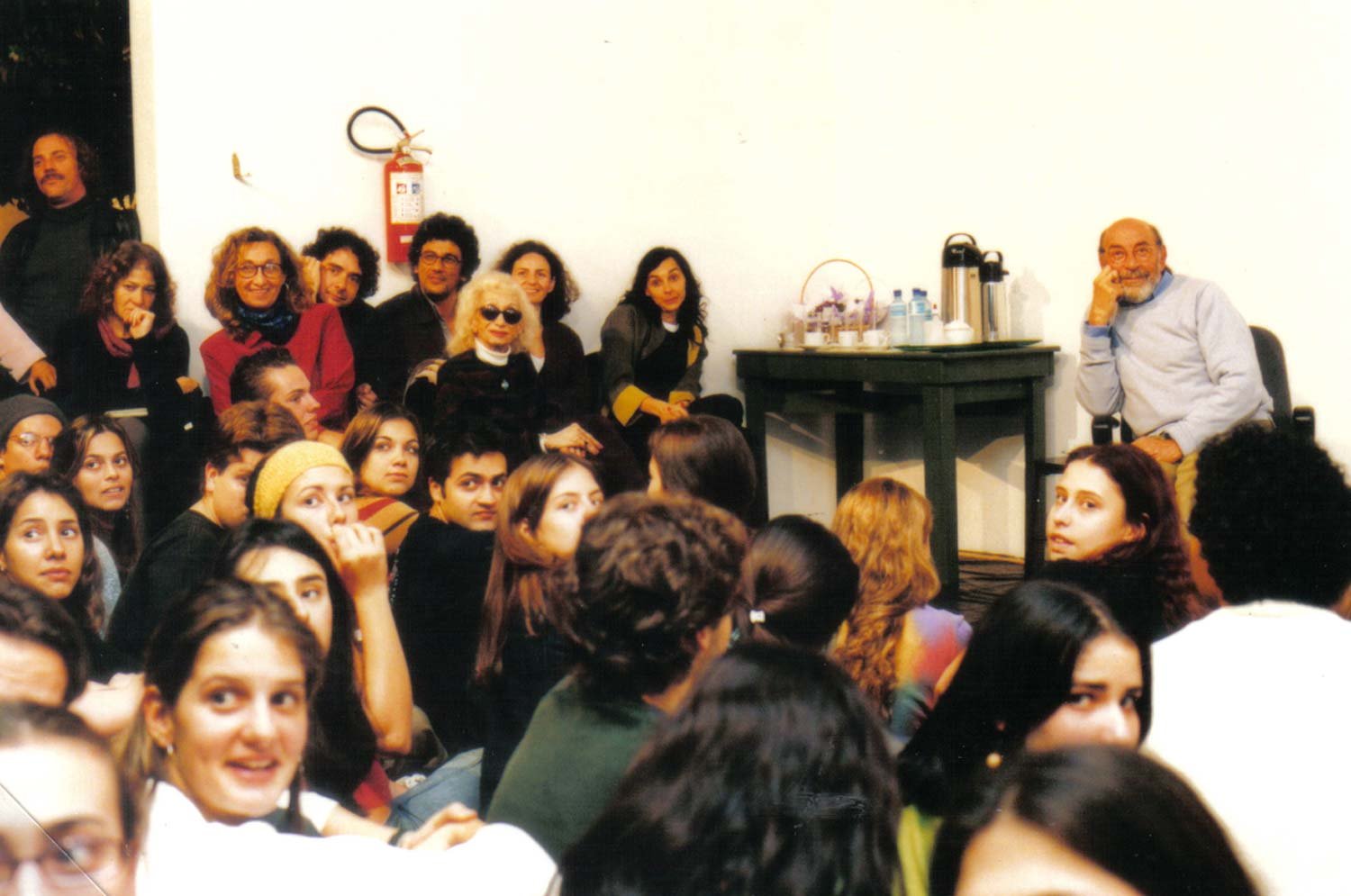 Vários jovens e alguns adultos sentados e em pé em uma sala, assistindo a uma apresentação ou conversa. Ao fundo, uma mesa com chaleira, copos e garrafas de água. Pessoas sentadas e observando atentamente.