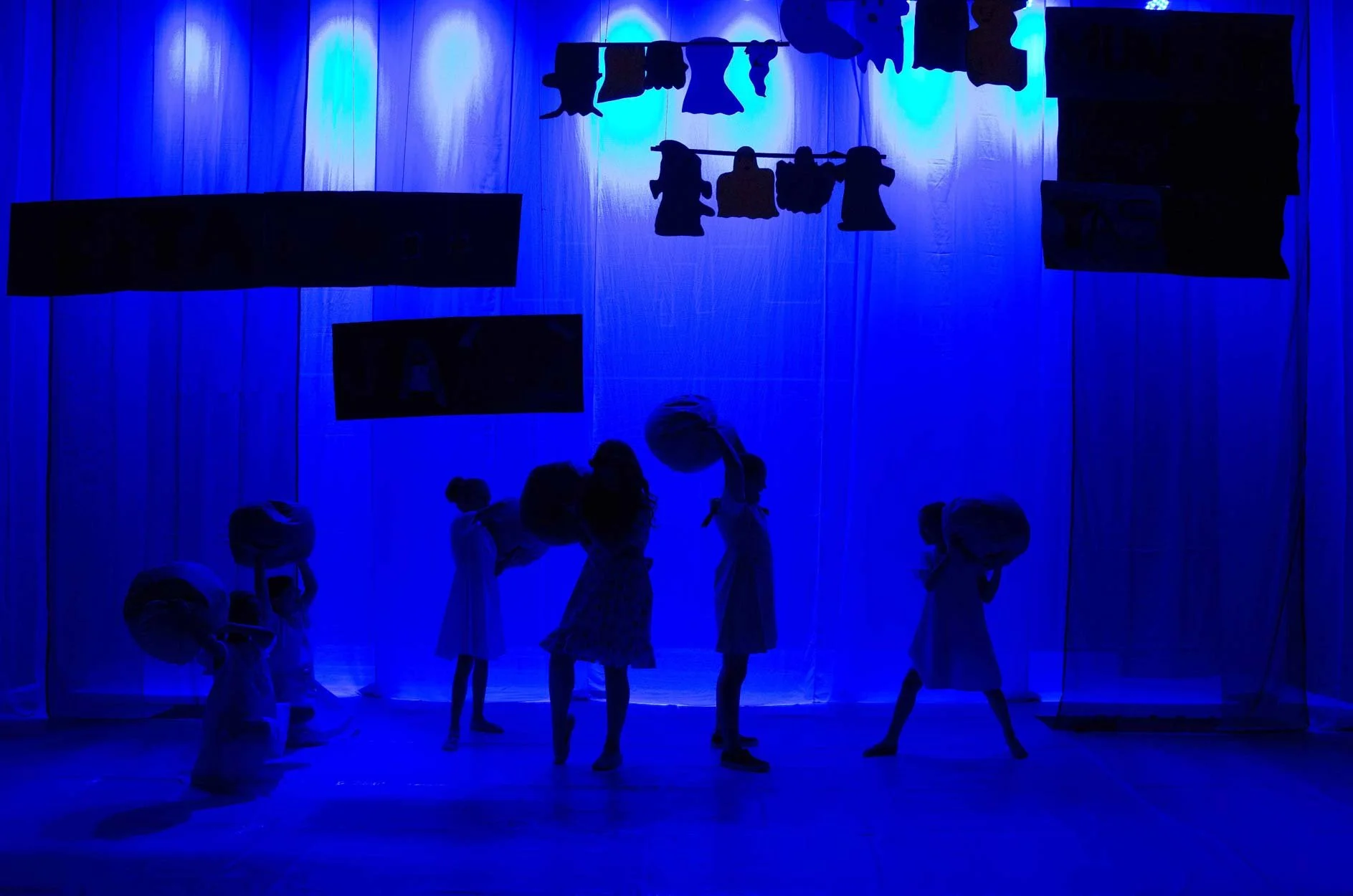 Crianças participando de uma apresentação de teatro com iluminação azul, vestidas com roupas claras, em um palco com pano de fundo branco e roupas penduradas acima.