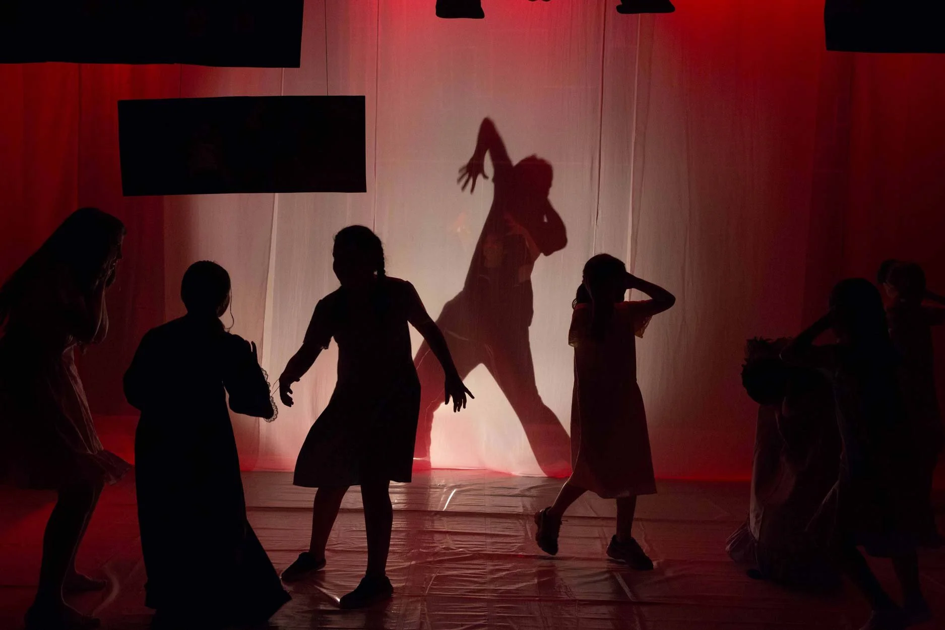 Silhuetas de crianças brincando em uma cortina de fundo iluminada com luz vermelha, com uma sombra de um cavalo projetada na cortina.
