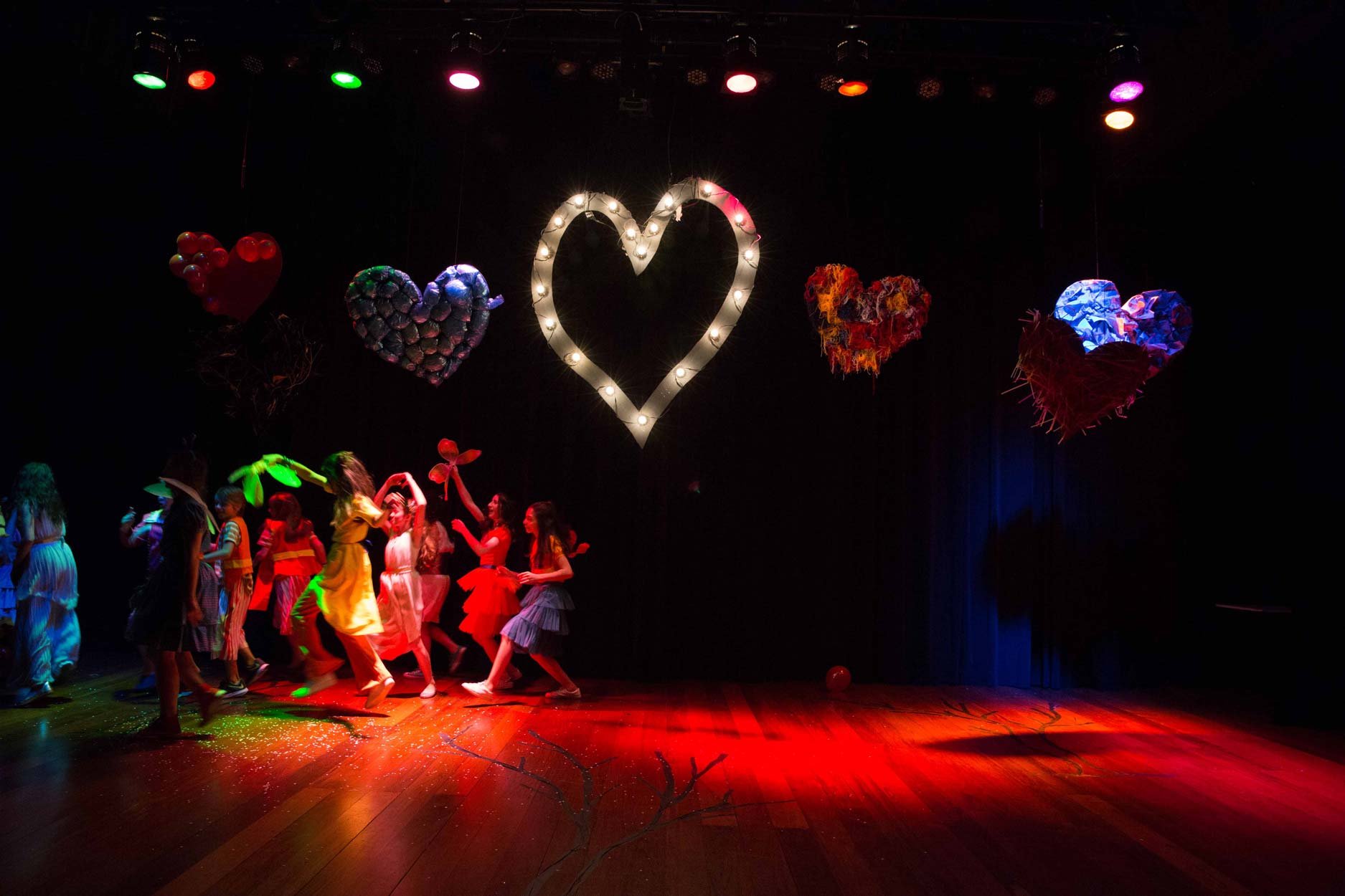 Crianças dançando em um palco decorado com corações iluminados e coloridos ao fundo.