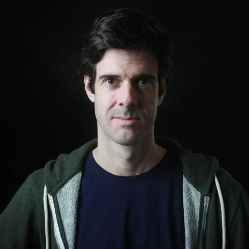 Retrato de um homem com cabelo escuro, expressão neutra, usando camisa azul e jaqueta verde escura, contra fundo preto.