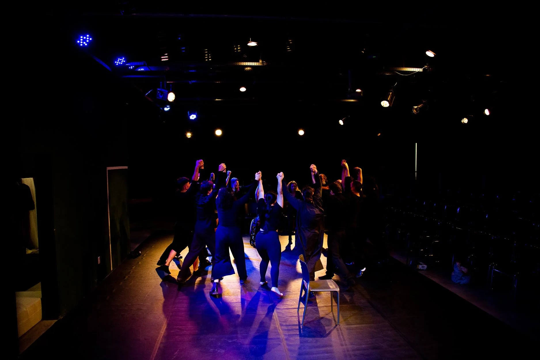 Grupo de pessoas no palco de teatro, com as mãos levantadas, sob iluminação de luzes de palco, enquanto uma cadeira está no centro do palco. Fundo escuro.