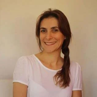 Mulher sorridente com cabelo castanho e pele clara, usando camiseta branca, em fundo claro.