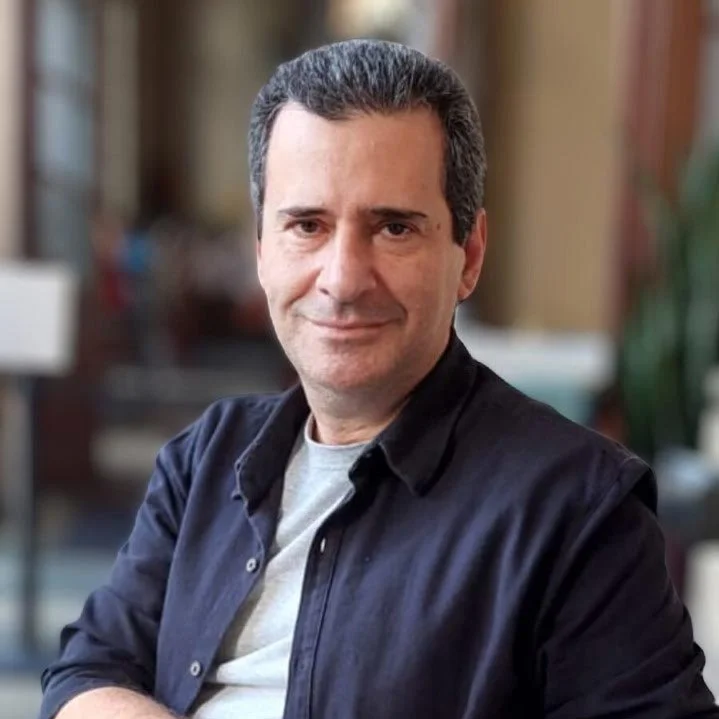 Retrato de um homem com cabelo escuro, pele clara, vestindo uma camisa azul escura, olhando para a câmera com expressão séria contra fundo preto.