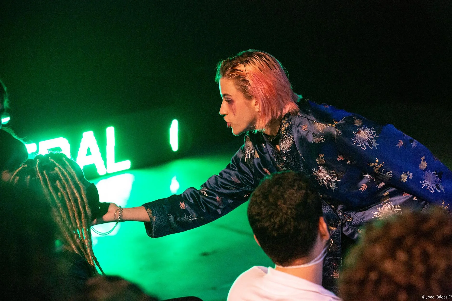 Pessoa com cabelo rosa, de perfil, oferece ajuda a uma pessoa de dreadlocks. No fundo, há letras verdes que parecem formar a palavra 'REAL'.