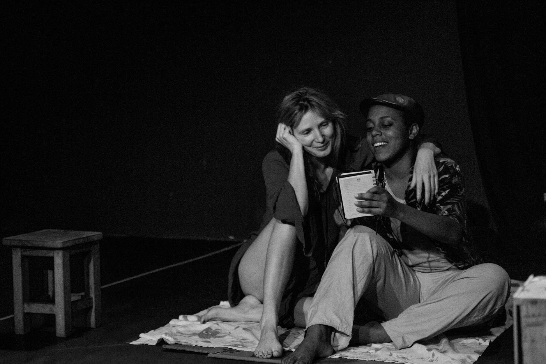 Duas pessoas sentadas em um tapete, uma delas segura um caderno, em um ambiente escuro.