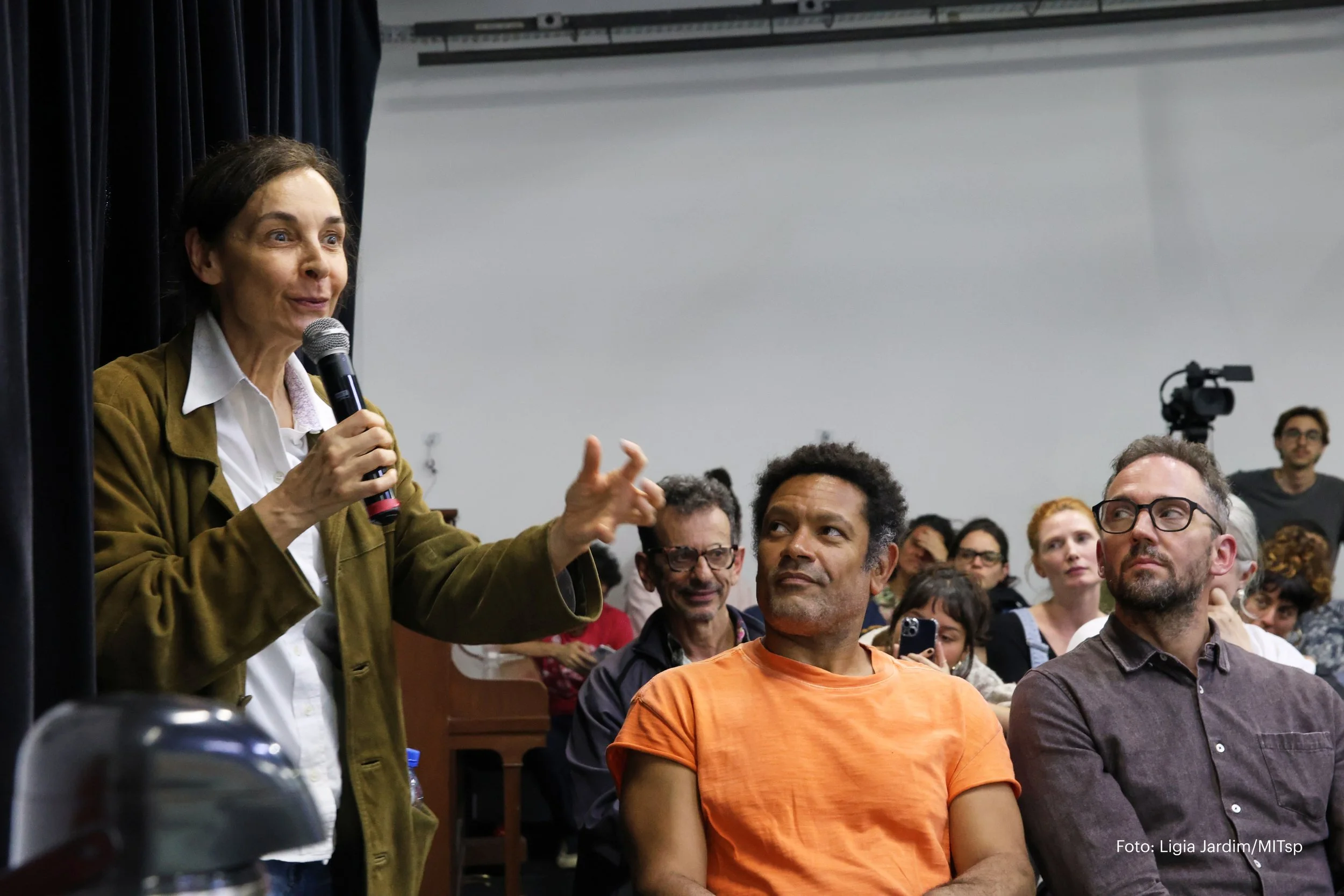 Mulher falando ao microfone em uma apresentação ou discussão, com um público atento e interessado ao redor, em um ambiente de sala de conferências ou palestra, com câmeras e equipamento de gravação no fundo.