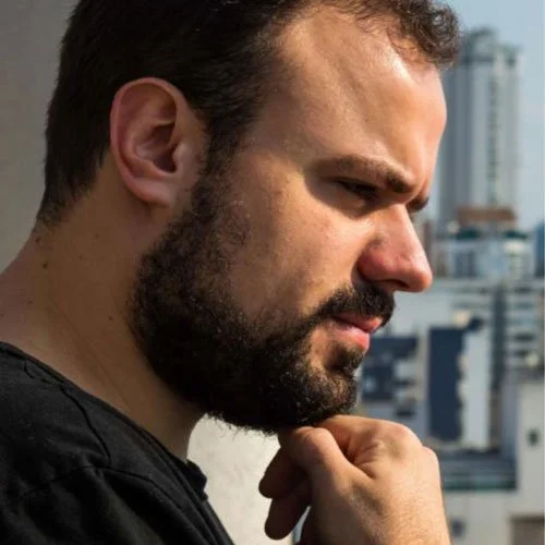 Homem com barba, cabelo castanho, olhando pensativo para o lado, com fundo de cidade com prédios