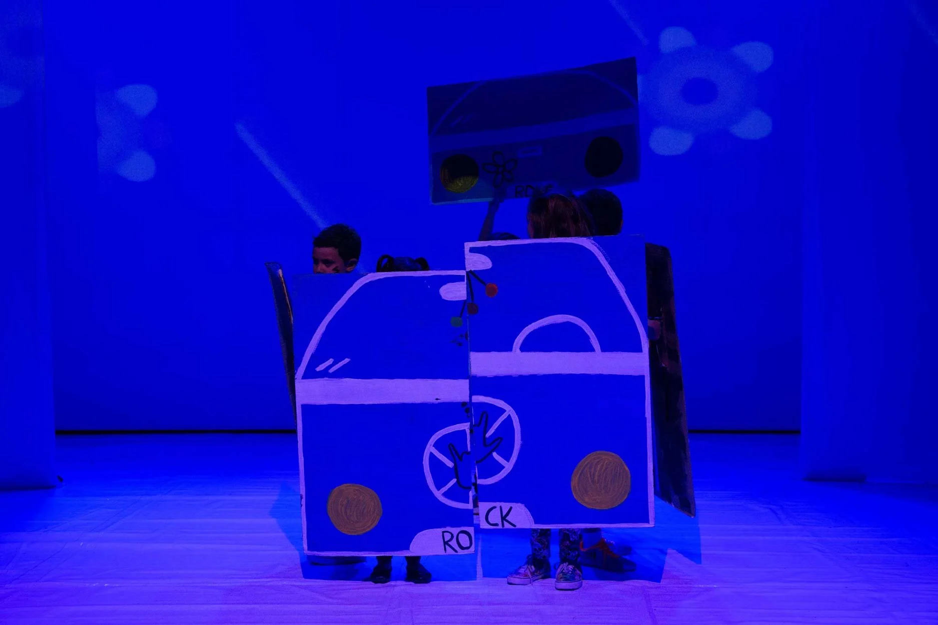 Crianças em uma apresentação teatral com adereços em estilo de carro e um painel de corrida, sob luz azul.