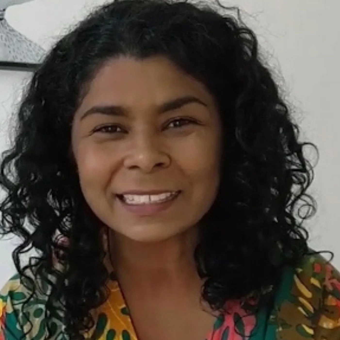 Mulher sorridente com cabelo cacheado preto, usando blusa colorida com padrões verdes, amarelos e vermelhos, em fundo branco.