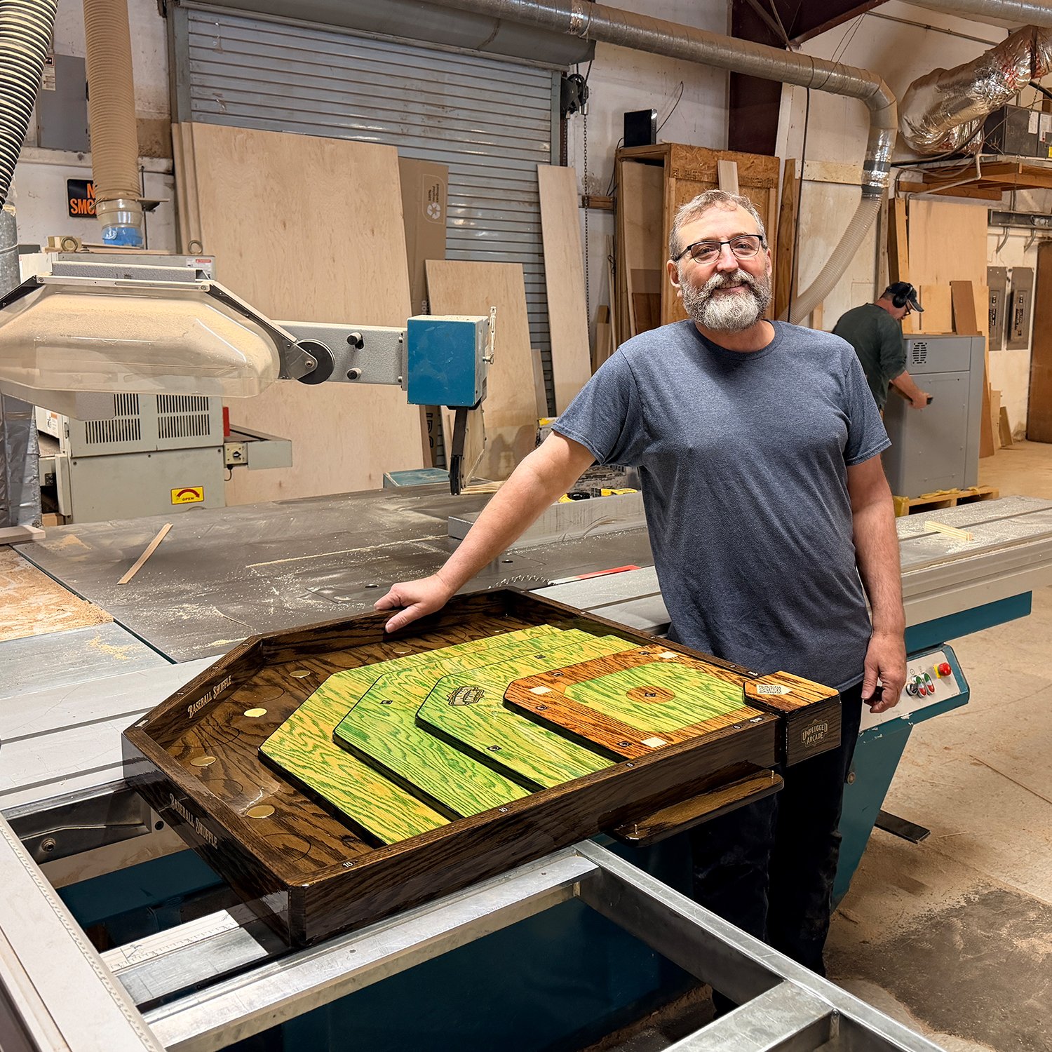 A man with glasses and a beard smiles, standing next to a table saw in a woodworking shop. He has one hand resting on a colorful, handcrafted wooden tray or game board on the table. In the background, another person is working near an electrical panel, with various wood panels and tools around.