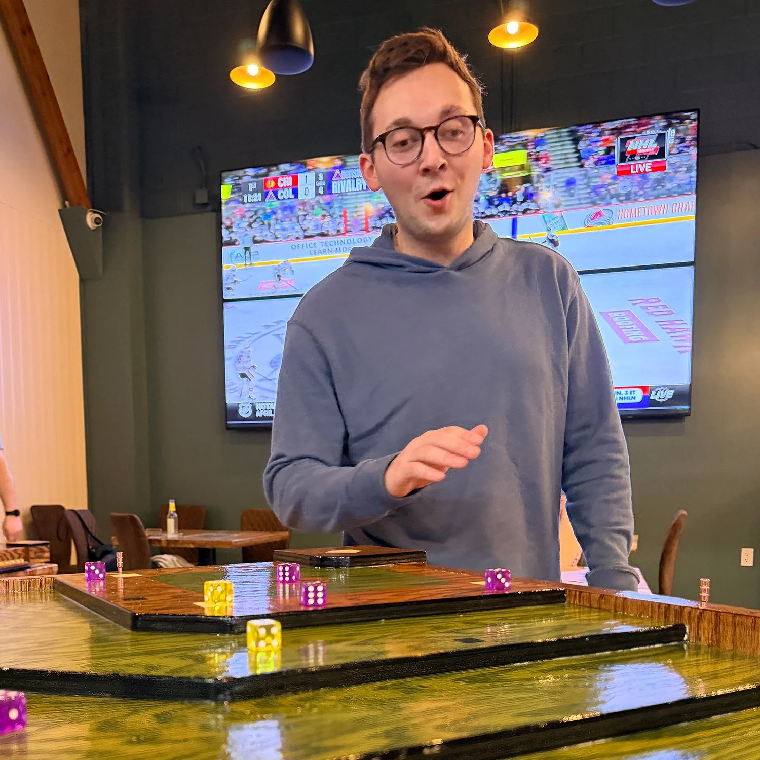 A player or group of friends are playing the dice-flicking party game, Baseball Shuffle, with drinks on the handcrafted oak game table. They are having a fun time at a brewery, taproom or social venue.
